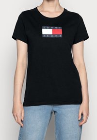 Svart bomulls t-shirt med en rektangulär logotyp med texten "TOMMY" och "JEANS" i vitt och röd på en blå bakgrund. Kort ärm, rund hals.