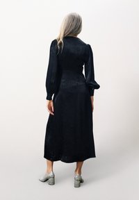 Schwarzes Satin-Kleid mit langen puffigen Ärmeln, strukturiertem Blumenmuster und fließender Silhouette. Kombiniert mit glänzenden silbernen Blockabsätzen.