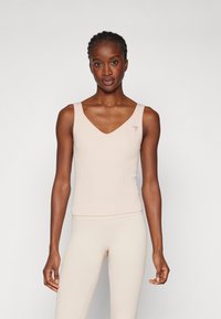 Ribbet tanktop i myk beige med V-hals og en liten triangulær logo på venstre bryst. Materialet ser lett og elastisk ut.