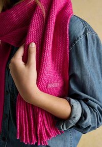 Jasnorůžová pletená šála s třásněmi, zdobená texturovaným vzorem a viditelnou značkovou cedulkou. Nošena přes džínovou košili.