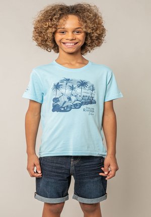Enfant souriant aux cheveux bouclés portant un t-shirt bleu clair avec un motif de trottinettes et de palmiers, et un short en denim foncé retroussé.