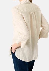 Lnian shirt w jasnym beżu z zaokrąglonym dołem, rękawami o długości trzech kwart i marszczeniem z tyłu. Kieszenie widoczne na ciemnoniebieskich spodniach.