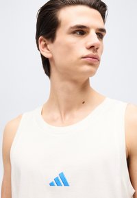 Νεαρός άνδρας με κοντά σκούρα μαλλιά, φορώντας λευκό αμάνικο μπλουζάκι με μπλε λογότυπο Adidas, κοιτάζει προς τα δεξιά σε απλό φόντο.