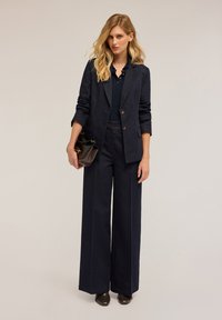 Námořnický modrý denimový oblek s dvouřadým blejzrem a širokými kalhotami. Blejzr má ohrnuté rukávy, doplněný černou kabelkou a tmavou obuví.