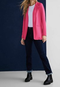 Nainen, joka on pukeutunut kirkkaan vaaleanpunaiseen bleiseriin valkoisen paidan, tummien, käärittyjen farkkujen ja mustien nilkkasaappaiden kanssa, seisoo kaksiväristä taustaa vasten.