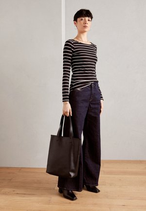 Gestreiftes Langarmoberteil in Schwarz und Weiß, kombiniert mit einer dunklen, weit geschnittenen Jeans. Das Model hält eine schwarze Ledertasche mit langen Griffen.