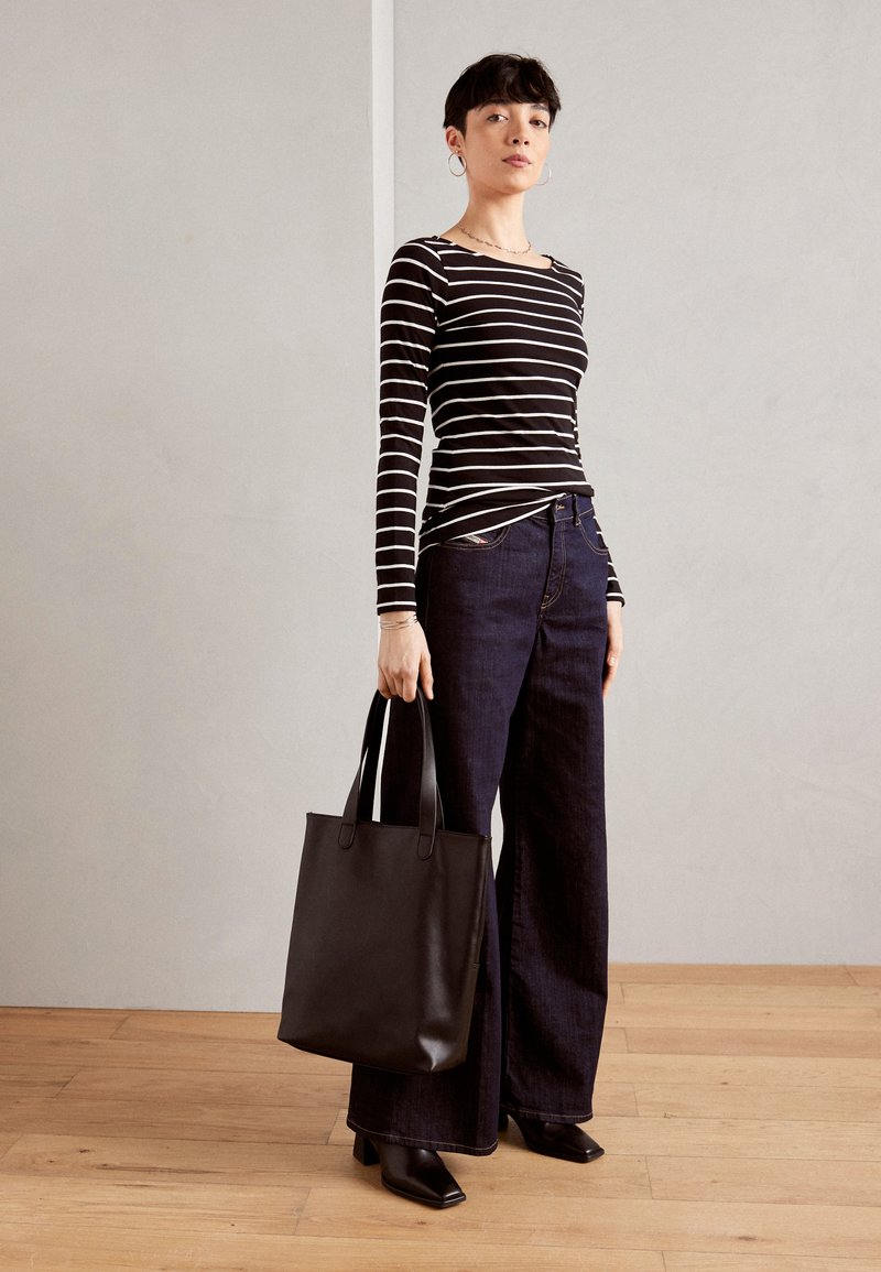Gestreiftes Langarmoberteil in Schwarz und Weiß, kombiniert mit einer dunklen, weit geschnittenen Jeans. Das Model hält eine schwarze Ledertasche mit langen Griffen.