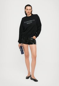 Felpa nera con stampa "CONCRETE BORN", abbinata a pantaloni corti neri lucidi. Il modello tiene una borsa blu con texture e indossa delle ballerine nere.