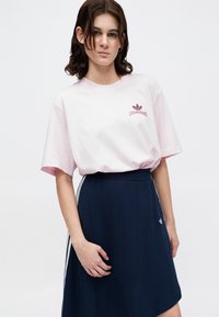 Jauns cilvēks ar vidēji gariem tumšiem matiem, uzvilcis gaiši rozā Adidas t-kreklu un tumši zilu svārku ar baltām svītrām.