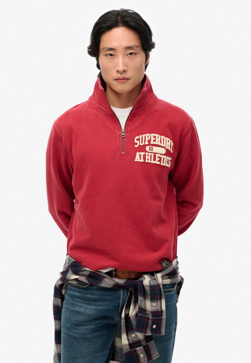 Sweatshirt zippé rouge avec "SUPERDRY ATHLETICS" imprimé en beige. Porté par-dessus une chemise blanche, associé à un jean bleu et une chemise à carreaux grise nouée autour de la taille.