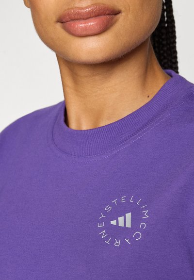 Lilla crewneck sweatshirt laget av bomull med et sirkulært hvitt logo som har tre striper og tekst i en moderne skrifttype.