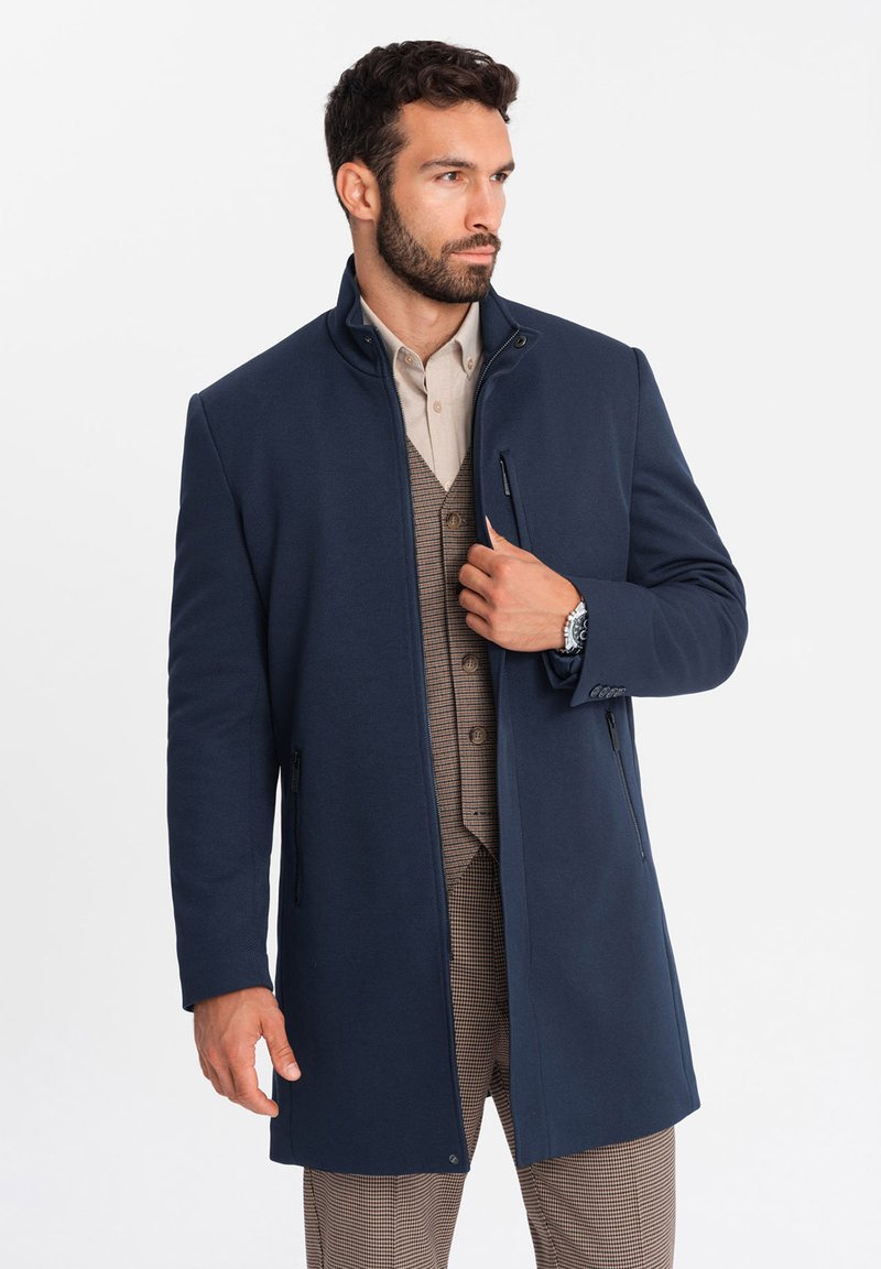 Cappotto blu navy con tasche con zip, colletto alto e vestibilità su misura, indossato sopra un gilet a fantasia, con un tessuto liscio e strutturato.
