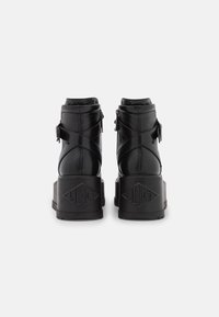 Botas negras de piel hasta el tobillo con una suela gruesa y texturizada, correas cruzadas y un detalle de hardware plateado en el costado. Diseño de tacón geométrico elegante.