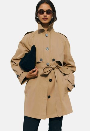 Frau trägt beigen Trenchcoat mit Gürtel und Sonnenbrille, hält eine schwarze Clutch und steht vor einfachem hellem Hintergrund.