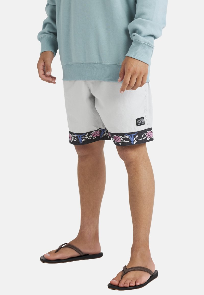 Man wearing light grey shorts with floral trim, teal sweatshirt, and brown flip-flops, standing on plain white background.