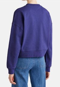 Fialová oversize mikina s kulatým výstřihem, žebrovanými manžetami a zkráceným lemem, v kombinaci se světle modrými džíny rovného střihu.