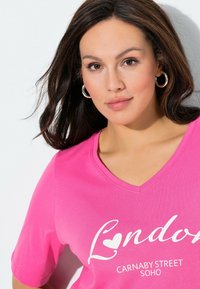 T-shirt rosa in cotone con scollo a V, caratterizzata da un motivo grafico bianco con la scritta "London Carnaby Street Soho" in un font giocoso.