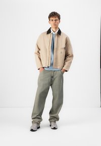 Beige Canvasjacke mit schwarzem Kragen, darunter ein blauer Pullover, lockere grüne Jeans und graue Sneakers. Mit Logopatch auf der Jacke.