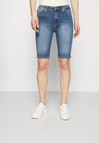 Džínové kraťasy středně modré barvy s pěti kapsami, ohrnutými lemy a zapínáním na zip, doplněné bílými teniskami.