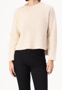 Suéter beige de punto con textura acanalada, cuello redondo y mangas largas, combinado con pantalones negros. Largo cropped con aberturas laterales.