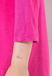 Top a maniche corte di un vivace colore rosa, realizzato in un tessuto morbido, con un dettaglio di bottone sulla manica e una leggera texture melangiata.