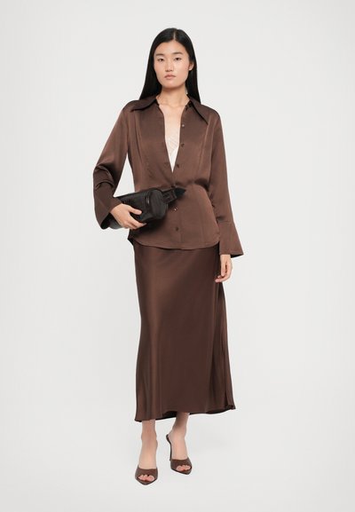 Brown satin long-sleeve shirt with buttons, paired with a matching satin skirt. Model holds a black textured handbag and wears brown open-toe heels.