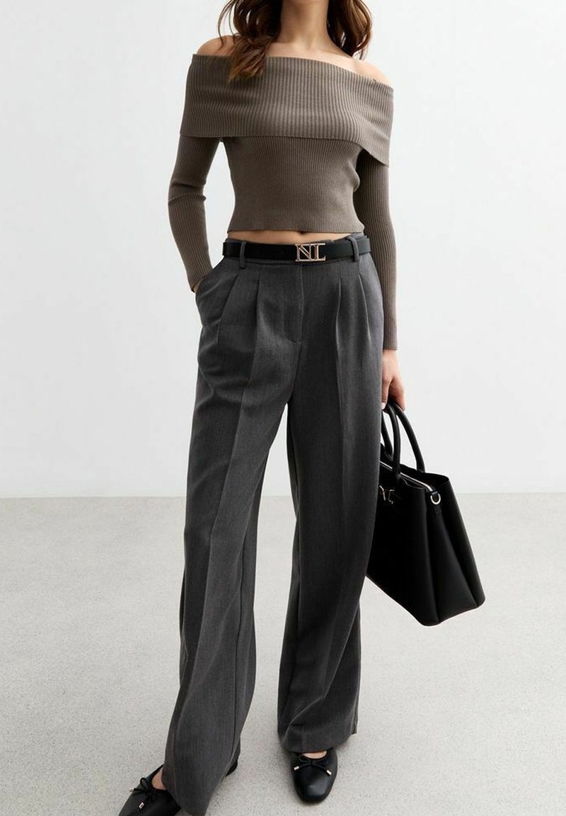 Femme portant un pull court taupe épaules dénudées, pantalon gris taille haute à jambes larges, ceinture noire, ballerines noires, tenant un sac à main noir.