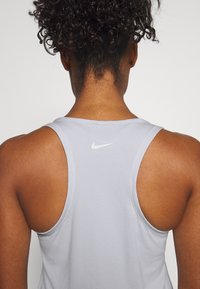 Femme aux cheveux bouclés portant un débardeur de sport Nike gris clair, vue de dos sur un fond uni.