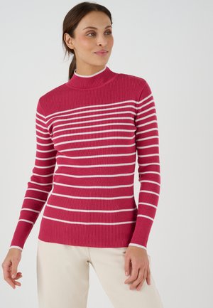 Vrouw in een rode geribbelde trui met witte horizontale strepen en lichte broek, staand tegen een egale achtergrond.
