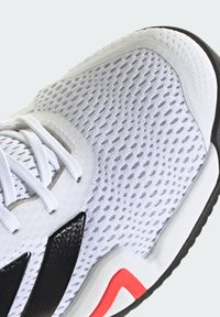 Bela športna obutev z zgornjim delom iz zračne mreže, črnimi poudarki, oblazinjenim ovratnikom in teksturirano gumijasto podplatom z rdečim detajlom blizu pete.