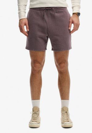 Homme portant un short à cordon violet foncé, des chaussettes blanches, des baskets montantes beiges et une chemise blanche à manches longues, debout les mains détendues.