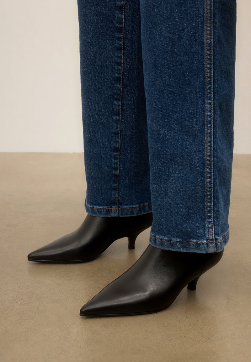 Pieds portant des bottines noires à bout pointu et talon kitten, associées à un jean droit bleu sur un sol beige.