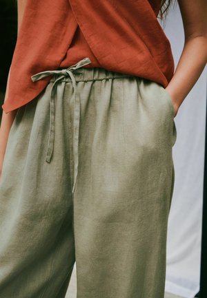 Løse olivengrønne linnedbukser med snøre i taljen, båret med rustfarvet top, hånd i lommen, beskåret udsigt fra taljen til midt på låret.