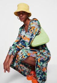 Vestido colorido e estampado com mangas longas, fenda até ao joelho, combinado com um chapéu estilo balde amarelo, uma bolsa tecida verde e sandálias de plataforma laranja.