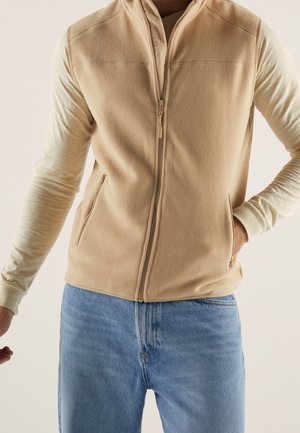 Homme portant un gilet en polaire beige zippé par-dessus une chemise crème à manches longues et un jean bleu, debout devant un fond clair.