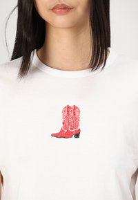 Valkoinen t-paita, jossa on punainen cowboy-boot-grafiikka, jossa on monimutkaisia valkoisia yksityiskohtia ja musta korko, sijoitettuna keskelle eteen.