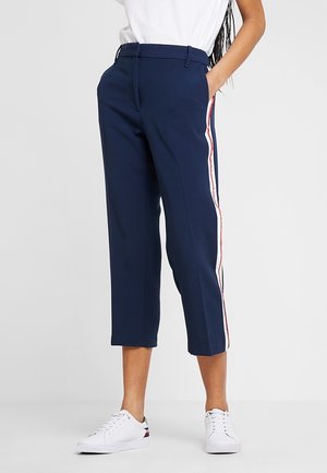 Woman wearing navy cropped pants with white and red side stripes, white sneakers, and a white tucked-in top, standing with one hand in pocket.