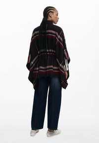 Schwarzer karierter Poncho mit burgunderfarbenen und weißen Akzenten, lockerer Schnitt, runder Ausschnitt und asymmetrischem Saum, kombiniert mit dunkelblauen Jeans und hellen Sneakers.