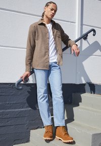 Chemise à carreaux marron à boutons sur un t-shirt blanc, jeans bleu clair, et bottes beige avec des lacets jaunes, debout sur des marches grises.