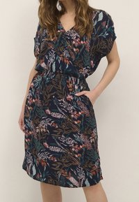 Femme portant une robe bleu marine mi-longue avec un motif floral coloré et des manches courtes, debout avec une main dans la poche.