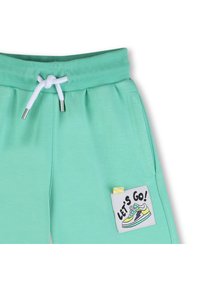 Πράσινο-μέντα φόρμας με κορδόνι στη μέση, πλαϊνές τσέπες και ένα έμπλαστρο στην αριστερή πλευρά με γραφικό αθλητικού παπουτσιού και το κείμενο "ΑΡΧΙΖΟΥΜΕ!".