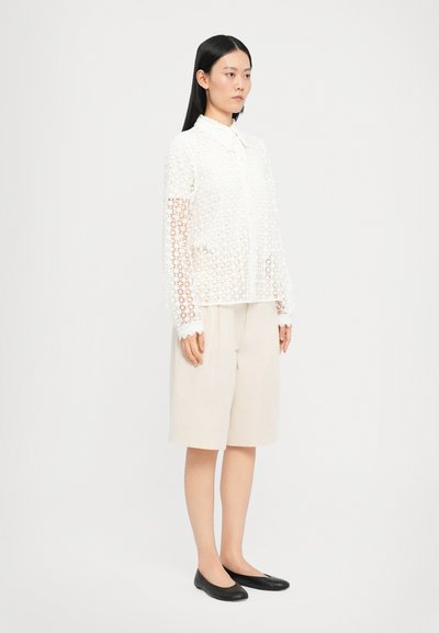 White sheer lace blouse with a collar and button closure, worn over beige shorts. Black flat shoes. Model stands against a plain background.