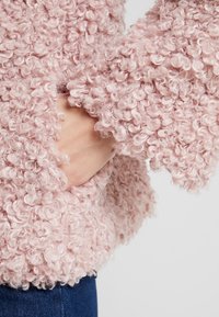 Chlupatý růžový svetr s texturovaným, kudrnatým materiálem. Má široké rukávy a kapsu, na obrázku je to oblečené s jeansy.