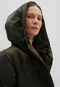 Donkergroene, puffy jas met een grote, verstelbare capuchon. Bevat een gladde textuur en kleine metalen sluitingen. Minimalistisch ontwerp.