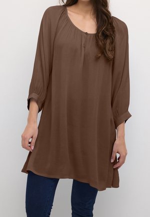 Kvinde iført løs, brun, langærmet tunika med knappet udskæring kombineret med mørkeblå jeans mod ensfarvet baggrund.