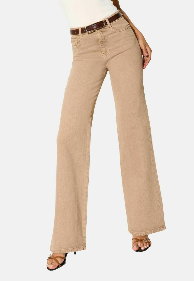 Beige højtaljede jeans med vide ben og brunt bælte, båret med brune åbne sandaler med stropper på en hvid baggrund.