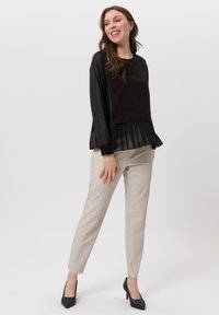 Vrouw die glimlacht, gekleed in een zwart langemouwtop met een doorzichtige geplooide onderrand, beige broek en zwarte hoge hakken, staande tegen een witte achtergrond.