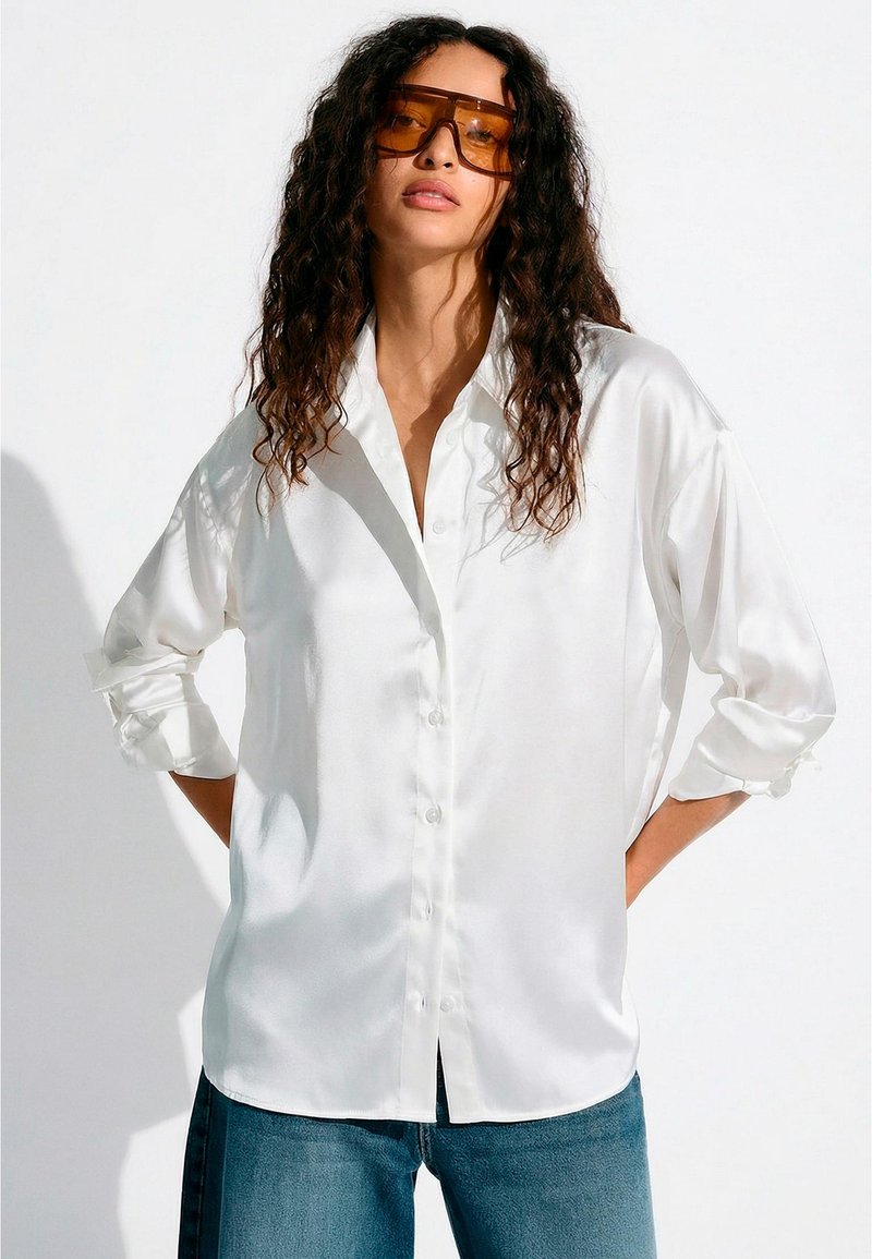 Göndör hajú nő túlméretezett, borostyánszínű napszemüveget visel, laza fehér szatén gombos ingben és kék farmerben, fehér háttér előtt.