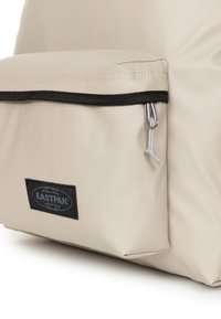 Borsa Eastpak beige con finitura texturizzata, dotata di una tasca frontale con zip nera e un patch con logo sul lato. Materiale resistente.
