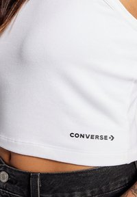 Top corto blanco hecho de algodón, con un logo negro de "CONVERSE" en la parte inferior derecha y un diseño simple y clásico.